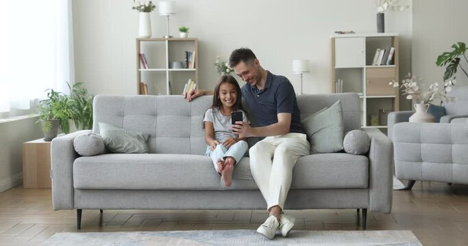 Cheerful preschool kid girl and happy dad taking funny video selfie on smartphone, resting on home sofa together, holding mobile phone, grimacing at screen, using media app for creative portraits