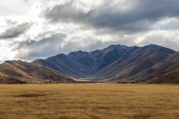 Photograph of a dry mountain range running behind a large brown agricultural field with low level grey clouds on the South Island of New Zealand - Powered by Adobe