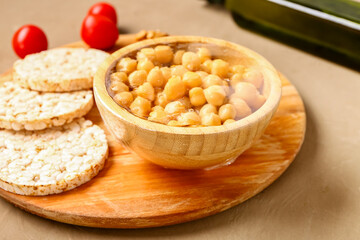 Bowl of fresh chickpeas covered with plastic food wrap and rice crackers on brown background