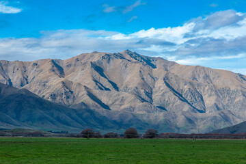 Fototapeta premium Photograph of a mountain range running behind a large green agricultural field with low level grey clouds on the South Island of New Zealand