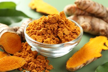 Aromatic turmeric powder and raw roots on green background, closeup