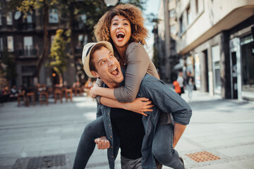 Fototapeta premium Young man carrying his girlfriend on his back while on a date in the city