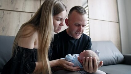 Caucasian new-made parents canoodling their newborn son. Parents sit on the sofa waving a baby and speaking to it.