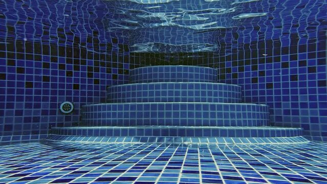 A woman walks along the bottom of the pool on her toes, climbs a ladder of blue tiles and comes out of the water. The sun's rays break through the ripples on the surface of the water.