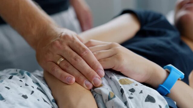 Male hand with wedding ring holding female one. Husband supporting pregnant wife lying in hospital bed. Blurred backdrop.