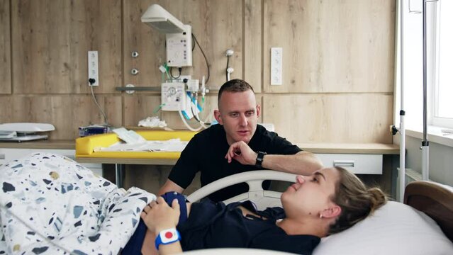 Man wearing black t-shirt talking to his wife lying in hospital bed. Woman is checked before baby delivery.