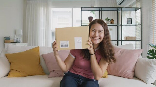 Young teen Gen Z girl asia people enjoy buy order gift postal parcel online shop. Happy receive courier carton box by fast express post mail omni channel retail store joy smile sitting at sofa home.