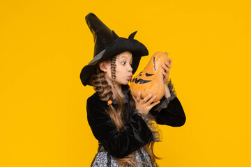 A little girl in a witch costume holds an evil pumpkin in her hands. Children's holiday carnival on Halloween. A happy child rejoices in a pointed black wizard hat. Yellow isolated background.