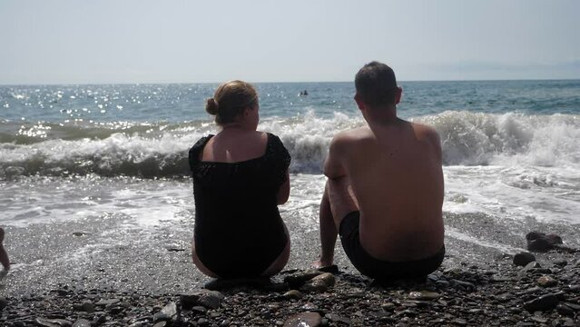 A young couple in swimsuits are sitting on a pebble beach with huge waves. Summer vacation at the sea. The family is sitting on the seashore in stormy weather. View from the back.