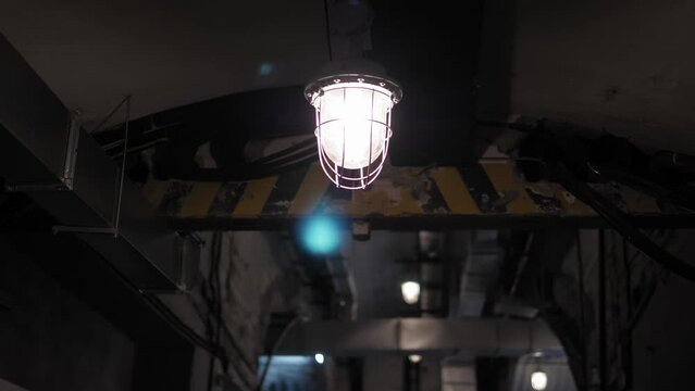 An explosion-proof lamp with a grille hangs from the ceiling in the tunnel of the bomb shelter. An empty scary dark corridor, a basement with concrete walls. Old electric lamps on the ceiling.