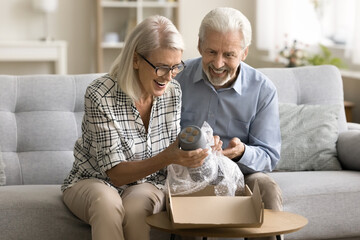 Cheerful senior online store customers couple opening parcel from webstore at home, taking object...