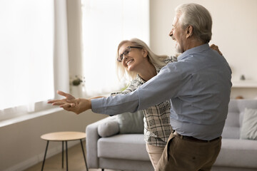 Joyful older retired husband and wife dancing to music, having fun, training pair social dance, holding outstretched hands, enjoying home party, activity, having fun, laughing