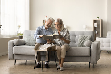 Happy senior married couple taking photo album from received open cardboard box, reviewing pictures, sitting, laughing, enjoying leisure in comfortable couch in cozy home interior