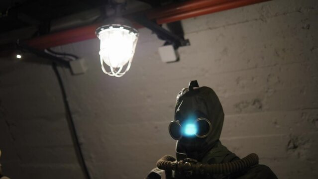 Close-up of a chemical protection suit and a gas mask on a mannequin in a dark tunnel or bomb shelter. An old electric lamp with a grille hangs on the ceiling. Nuclear war.