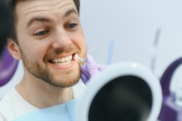 Dentist in blue medical gloves applying sample from tooth enamel scale to smiling man patient teeth to pick up right shade, teeth bleaching procedure - obrazy, fototapety, plakaty