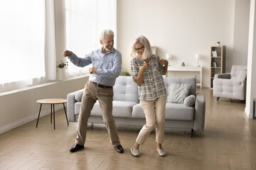 Cheerful active retired elder dancing to disco music in spacious living room, having fun, enjoying sportive hobby, home party, celebrating anniversary, family special date
