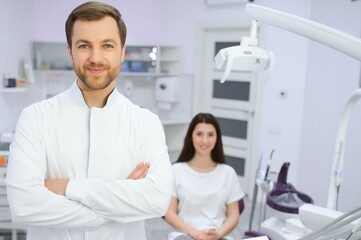 Portrait of positive male dentist with crossed hands posing at camera with patient and nurse on background