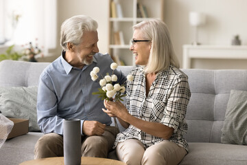 Happy senior couple celebrating wedding day anniversary, sitting on home couch with flowers, smiling, laughing. Positive elder husband giving bouquet to cheerful mature wife