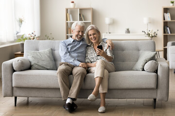 Positive retired elderly grandpa and grandma enjoying wireless Internet technology on home couch together, using smartphone, talking on video call, resting on sofa in cozy interior