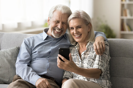 Happy senior retired couple of grandparents talking on online video call, using mobile phone on home sofa for remote Internet communication, enjoying conversation, family chat