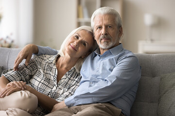 Confident married retired senior couple in love posing on home sofa for family portrait together, sitting close, hugging with trust, affection, looking at camera with smiling and serious face