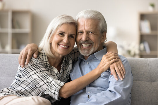 Happy attractive older retired couple in love posing for family portrait, hugging on home couch, looking at camera with perfect toothy smiles. Cheerful senior husband and wife video call shot