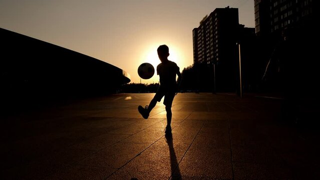 The silhouette of a little boy stuffing a ball in a city park in summer at sunset. The child is engaged in football. 