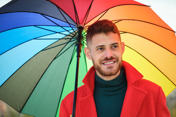 Portrait of latin young man with a red coat and a rainbow umbrella in the street in autumn.