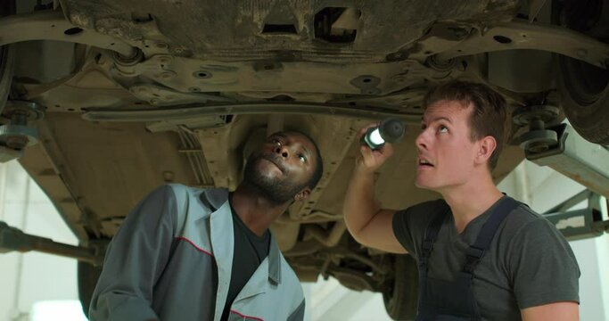 Two young hardworking auto mechanics working on undercarriage of car, two mixed-race guys examing technical condition of the lifted broken car. automotive car repair concept