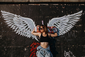 Young street-stylish beautiful girl with skateboard	and wings