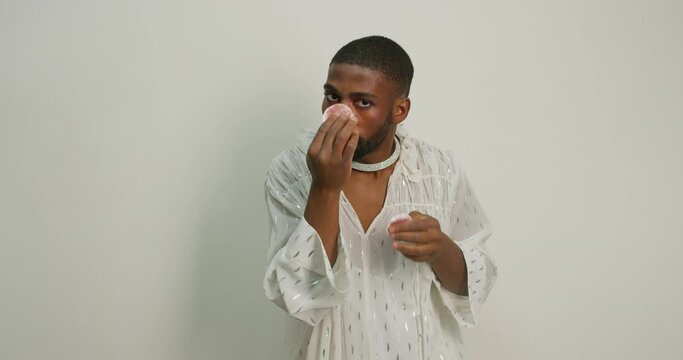 black man in fashionable outfit concentated on wiping cosmetics from face, trangender take care of face Slow motion Beauty, gay using cotton pad in studio with white wall