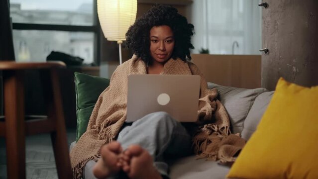 Pretty young woman freelancer under the blanket while working online typing browsing on laptop laying on sofa at late evening indoors Positive female enjoying great working day at cozy home