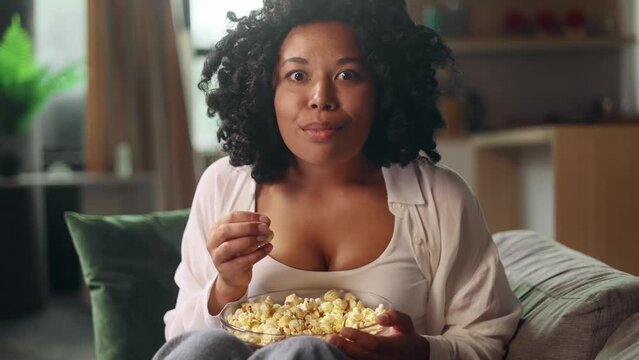 Camera view of addicted focused young curly woman watching interesting TV program film movie and eating popcorn alone at home Enchanted curly girl cannot take her eyes off the screen indoors