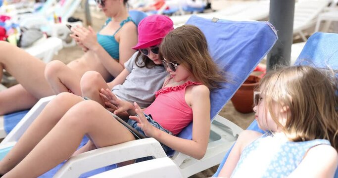 Teenage sisters lie on sun loungers playing game on tablet near mother. Technology and relaxation on beach with family concept slow motion
