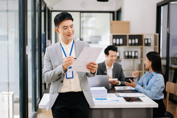 Young attractive Asian male office worker business suits smiling at camera with working notepad, tablet and laptop documents in office