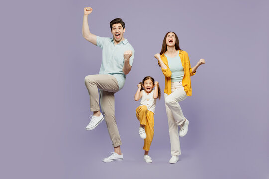 Full body young parent mom dad with child kid daughter girl 6 years old wear blue yellow casual clothes do winner gesture celebrate raise up leg isolated on plain purple background Family day concept