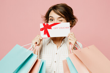 Young woman wears casual clothes hold shopping paper package bags cover mouth with gift coupon voucher card for store wink isolated on plain pink color background. Black Friday sale buy day concept.