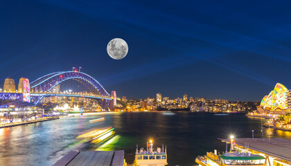 Obraz premium Panoramic night view of Sydney Harbour and City Skyline with a Sturgeon full Moon NSW Australia bright neon lights reflecting off the water