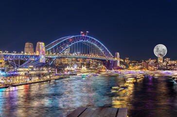 Obraz premium Panoramic night view of Sydney Harbour and City Skyline with a Sturgeon full Moon NSW Australia bright neon lights reflecting off the water