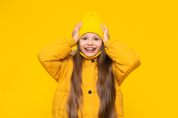 A little girl in a jacket and hat holds her head and smiles happily. A child in autumn,...