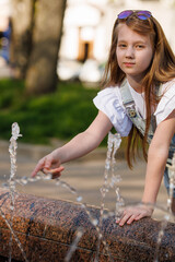 A little girl with blond long flowing hair and sunglasses on her head, in a denim overalls, white golfs and sneakers, a walk around the city with fountains