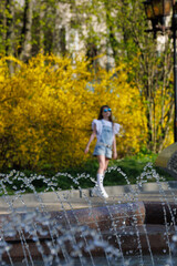 A little girl with blond long flowing hair and sunglasses on her head, in a denim overalls, white golfs and sneakers, a walk around the city with fountains