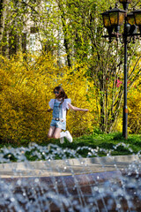 A little girl with blond long flowing hair and sunglasses on her head, in a denim overalls, white golfs and sneakers, a walk around the city with fountains