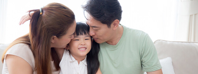 Happy asian family father and mother kiss cheek daughter sitting on sofa in living room at home,...