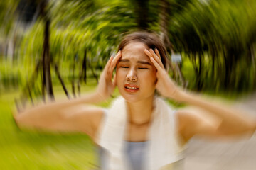 Asian woman exercising outdoors with discomfort hands gently massaging her head in too hot weather...