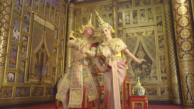Thai classic masked man from the Ramakien character with red mask dance with beautiful Asian woman wear Thai traditional dress and stay in front of Thai painting on public place wall.