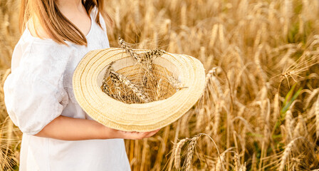 Happy girl in white dress with straw hat full of ears of wheat, rye, barley walking in yellow,...