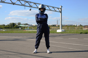 Woman racer in an outfit and a helmet stands in front of a full-length karting track before the start of the race with crossed arms