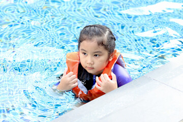 Fototapeta na wymiar Cute Asian girl with life jacket swimming in blue water, kid playing outdoor activity and leaning to swimming in the pool on a sunny day, having fun on summer vacation holiday weekend.