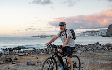 Happy active senior cyclist man at sea at sunset light with electric bicycle running on the beach - elderly man with helmet enjoying healthy lifestyle and freedom